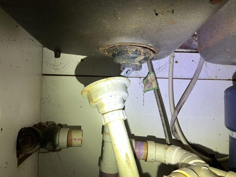 # Kitchen Sink Leak Repair

Responded to a water leak originating from under the kitchen sink and dripping into the basement below. Upon arrival, performed thorough inspection of the under-sink plumbing assembly, which showed signs of aging with yellowed and stained PVC components.

**Assessment:**
Examined the complete drain assembly including the metal sink drain flange, white plastic P-trap system, and all associated connections. Inspected the garbage disposal unit, flexible braided water supply lines, and vertical tailpiece connections. Identified source of leak at deteriorated drain connections showing visible discoloration and accumulated grime from months of slow seepage.

**Work Performed:**
- Disassembled affected sections of the P-trap assembly
- Cleaned and inspected all drain connections and sealing surfaces
- Replaced worn gaskets and compression fittings at leak points
- Reinstalled P-trap with proper alignment and secure connections
- Verified garbage disposal mounting a