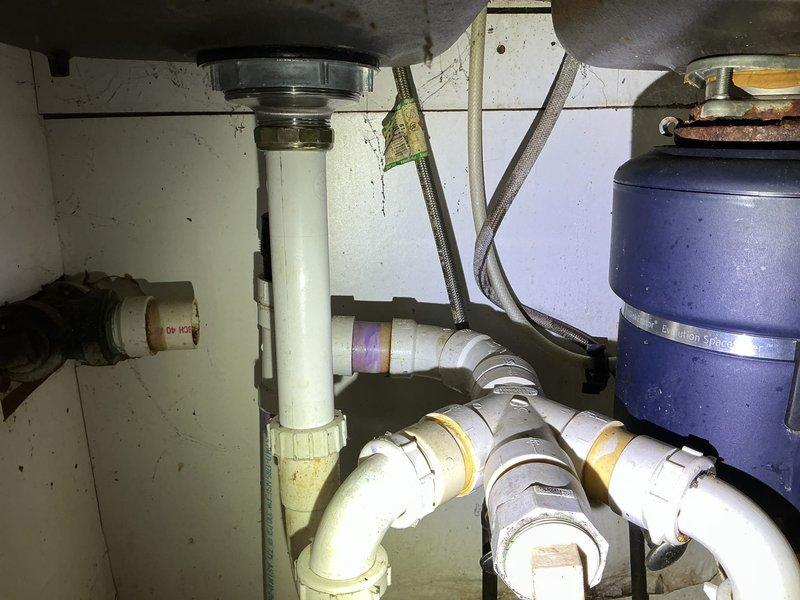# Kitchen Sink Leak Repair

Responded to a water leak originating from under the kitchen sink and dripping into the basement below. Upon arrival, performed thorough inspection of the under-sink plumbing assembly, which showed signs of aging with yellowed and stained PVC components.

**Assessment:**
Examined the complete drain assembly including the metal sink drain flange, white plastic P-trap system, and all associated connections. Inspected the garbage disposal unit, flexible braided water supply lines, and vertical tailpiece connections. Identified source of leak at deteriorated drain connections showing visible discoloration and accumulated grime from months of slow seepage.

**Work Performed:**
- Disassembled affected sections of the P-trap assembly
- Cleaned and inspected all drain connections and sealing surfaces
- Replaced worn gaskets and compression fittings at leak points
- Reinstalled P-trap with proper alignment and secure connections
- Verified garbage disposal mounting a