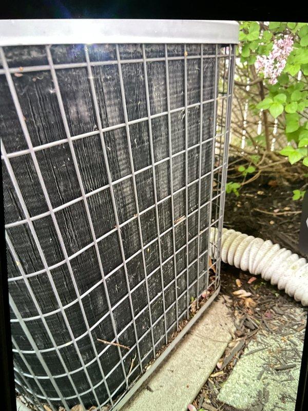 Completed Gold AC Inspection on 25-year-old residential air conditioning system with basement-located air handler. Upon arrival, performed comprehensive visual assessment of outdoor condenser unit, which revealed substantial accumulation of dirt, dust, and debris on cooling fins and heat exchanger surfaces, indicating extended period without maintenance. Condenser coils displayed heavy contamination with darkened, grayish-black appearance throughout visible fin surface area. Noted organic debris including dried leaves and twigs accumulated around unit base and caught in protective metal grille. Documented unit condition through photographic inspection, capturing extent of environmental contamination affecting heat transfer efficiency. Verified equipment operational status with homeowner prior to inspection. Explained weather-dependent inspection protocols regarding temperature and precipitation limitations for optimal assessment conditions. Unit protected by white metal cage grille in 