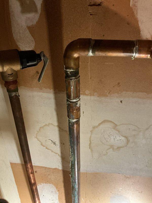 Responded to service call for leaking hot water supply line on commercial water heater serving six apartment units located in mechanical room. Upon arrival, inspected copper supply piping and identified active leak at corroded connection point near base of vertical supply line. Copper piping exhibited significant oxidation, green patina buildup, and corrosion damage consistent with long-term moisture exposure and age-related deterioration. Wall surfaces and flooring around piping showed evidence of water damage and staining from ongoing leak. Shut down water supply to affected heater to prevent further water damage. Assessed extent of corrosion on vertical copper runs and determined compromised pipe sections required replacement to restore system integrity. Documented existing conditions including corroded pipe sections, deteriorated fittings, and water-damaged surrounding areas for repair recommendations. Provided assessment that hot water supply piping system requires replacement of 