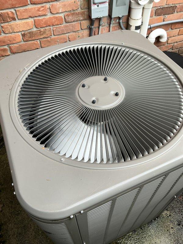Completed comprehensive Gold AC inspection on single residential split system with basement air handler and outdoor condenser unit, less than 10 years old. Performed visual assessment of outdoor condenser positioned on concrete pad against brick wall, confirming proper clearance and mounting. Inspected fan assembly through top grille, noting clean radial fins and well-maintained condition. Verified refrigerant lines and electrical connections at exterior unit, observing proper routing of gray flexible conduit along brick wall. Documented unit showing normal outdoor weathering with functional components and no visible damage. System confirmed operational at time of inspection with homeowner present.