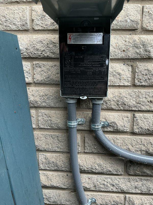 Completed comprehensive Gold AC Inspection on outdoor air conditioning unit located on exterior patio. Performed full system tune-up including inspection of electrical disconnect box, conduit connections, and unit housing. Verified proper operation of all components, checked electrical connections at disconnect panel, and documented equipment condition with photographic records showing disconnect box in open position with visible specifications panel and PVC conduit routing along exterior wall. Unit appears to be in functional condition with all exterior components properly secured and maintained.