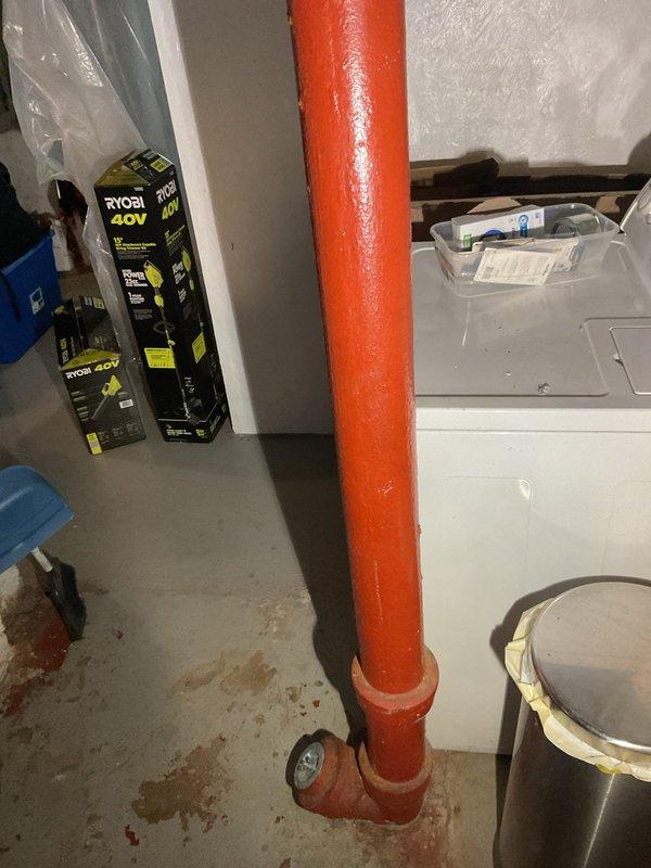 Responded to service call for deteriorating cast iron waste stack piping in basement utility area. Upon inspection, found vertical drain pipe with severely compromised structural integrity, exhibiting extensive surface corrosion, paint failure, and exposed metal substrate throughout the length of the stack. The cast iron piping showed advanced degradation consistent with age-related deterioration, with visible rust staining on surrounding concrete floor indicating potential historical leakage or seepage. Documented existing conditions through photographic evidence showing the full extent of pipe deterioration from ceiling to floor penetration, including flared hub connections and compromised protective coating. Assessed replacement requirements for complete staff piping system upgrade from obsolete cast iron to modern piping materials to prevent future failure and ensure code compliance.
