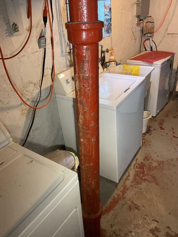 Responded to service call for deteriorating cast iron waste stack piping in basement utility area. Upon inspection, found vertical drain pipe with severely compromised structural integrity, exhibiting extensive surface corrosion, paint failure, and exposed metal substrate throughout the length of the stack. The cast iron piping showed advanced degradation consistent with age-related deterioration, with visible rust staining on surrounding concrete floor indicating potential historical leakage or seepage. Documented existing conditions through photographic evidence showing the full extent of pipe deterioration from ceiling to floor penetration, including flared hub connections and compromised protective coating. Assessed replacement requirements for complete staff piping system upgrade from obsolete cast iron to modern piping materials to prevent future failure and ensure code compliance.