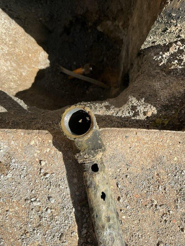 Responded to service call for reported gas leak beneath natural gas grill. Upon inspection, located corroded galvanized gas supply line in underground utility access area showing significant deterioration with visible perforations and rust damage along pipe exterior. Traced gas line from underground chamber to above-ground connection point at wooden post where shutoff valve and brass fittings were installed. Performed pressure test on gas system to verify leak location and assess system integrity. Documented extensive corrosion on underground pipe section requiring replacement to ensure safe operation. Shut off gas supply at valve with red handle to secure system until repairs could be completed. Recommended immediate replacement of compromised underground gas line section due to safety concerns from multiple perforation points and advanced pipe deterioration.