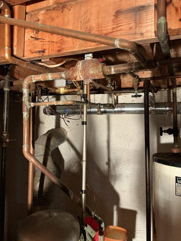 Provided estimate for complete removal of existing boiler piping system and capping of incoming water supply line to decommissioned boiler. Assessed extensive network of aged copper piping throughout basement mechanical room showing significant corrosion, green patina buildup, and oxidation on valve assemblies. Evaluated corroded red-orange valve components in upper section requiring removal. Inspected PVC drain configuration and black supply lines in utility area. Documented deteriorated plumbing infrastructure, water-damaged wooden surfaces, and accumulated debris. Estimate includes disconnection and removal of all copper pipes connected to boiler system, proper capping and securing of main water supply line to prevent future water flow to unit, and disposal of removed materials.