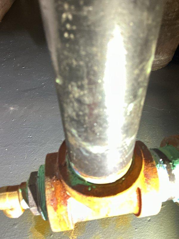 Responded to service call for boiler leaking onto floor from recent installation completed in March. Upon inspection, identified active leak at pipe connection point with visible water damage and significant corrosion present on brass fitting and galvanized piping. Photographic documentation captured corroded brass fitting with orange-brown oxidation staining at connection interface, green gasket seal, and deteriorated metal pipe showing heavy rust, green patina, and surface corrosion throughout vertical run. Confirmed leaking components were part of original installation work, requiring recall evaluation and repair of faulty plumbing connections.