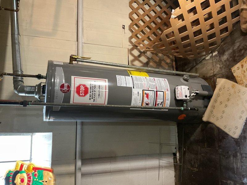 Responded to water heater leak in basement of 80-year-old residence. Upon inspection, found 13-year-old residential water heater with active profuse leaking from tank unit. Assessed piping connections, flue vent system, and surrounding plumbing infrastructure including exposed copper and black iron pipes with shut-off valves. Documented condition of water heater with manufacturer labels visible, noting unit positioning on concrete floor with containment mat. Evaluated ceiling-mounted supply and return lines with insulated piping and confirmed leak was isolated to basement area with no water migration to other floors. Provided assessment and recommendations for water heater replacement due to age of unit and severity of tank failure.