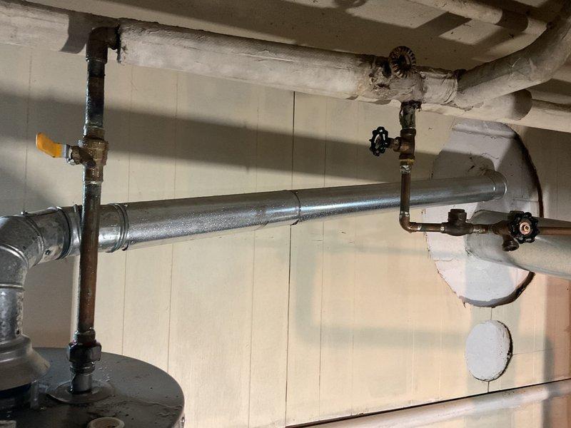 Responded to water heater leak in basement of 80-year-old residence. Upon inspection, found 13-year-old residential water heater with active profuse leaking from tank unit. Assessed piping connections, flue vent system, and surrounding plumbing infrastructure including exposed copper and black iron pipes with shut-off valves. Documented condition of water heater with manufacturer labels visible, noting unit positioning on concrete floor with containment mat. Evaluated ceiling-mounted supply and return lines with insulated piping and confirmed leak was isolated to basement area with no water migration to other floors. Provided assessment and recommendations for water heater replacement due to age of unit and severity of tank failure.
