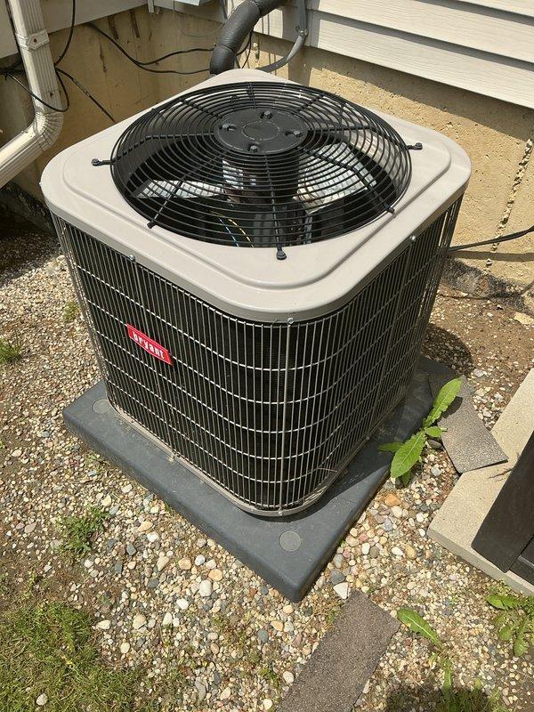 Completed comprehensive NSP AC inspection on a 5-year-old single-unit residential air conditioning system with air handler located in basement. Performed visual inspection of outdoor condensing unit installed on composite pad against exterior wall, verified proper installation with electrical disconnect box and refrigerant lines in place. Unit operational at time of inspection with no immediate service concerns identified. Documented equipment condition with photographs showing condenser on gravel base with adequate clearance, though minor vegetation growth noted around unit perimeter that may require periodic maintenance for optimal airflow.