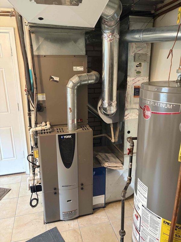 Completed comprehensive Gold AC inspection on 20-year-old York split system with air handler located in basement utility room and condenser unit positioned on exterior concrete pad. Performed full system evaluation including visual inspection of indoor air handler, ductwork connections, refrigerant line set, outdoor condenser unit with horizontal louvers and top grilling, electrical disconnect box, and copper refrigerant piping penetration through exterior wall. Verified operational status of all components, checked for proper airflow, inspected condensate drainage, examined electrical connections and conduit routing, and assessed overall system condition noting age of equipment at two decades of service.
