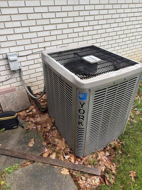Completed comprehensive Gold AC inspection on 20-year-old York split system with air handler located in basement utility room and condenser unit positioned on exterior concrete pad. Performed full system evaluation including visual inspection of indoor air handler, ductwork connections, refrigerant line set, outdoor condenser unit with horizontal louvers and top grilling, electrical disconnect box, and copper refrigerant piping penetration through exterior wall. Verified operational status of all components, checked for proper airflow, inspected condensate drainage, examined electrical connections and conduit routing, and assessed overall system condition noting age of equipment at two decades of service.