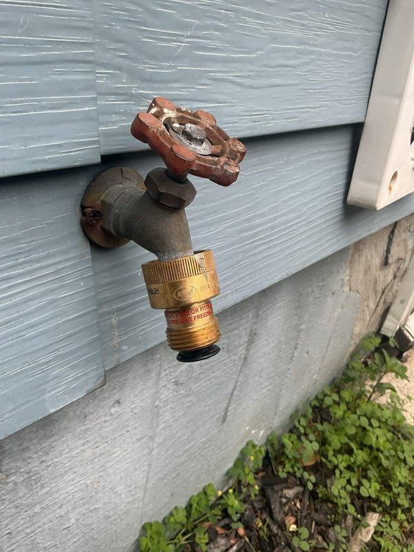Responded to service call for installation of vacuum breaker on sump pump discharge line to meet municipal code requirements. Upon arrival, located exterior sump pump discharge point on building's exterior wall with light blue vinyl siding. Existing discharge configuration consisted of copper pipe penetration through foundation with basic hose bib valve assembly.

Installed atmospheric vacuum breaker assembly on the discharge line to prevent potential backflow contamination of potable water supply. The vacuum breaker was mounted on tan square backing plate secured to exterior wall above the existing valve assembly. This device provides required air gap protection by allowing air to enter the system when pressure drops, preventing siphonage back into the building's water supply system.

Verified proper installation orientation with vacuum breaker positioned vertically and installed downstream of any shutoff valves per plumbing code standards. Tested valve operation and confirmed vacuum 