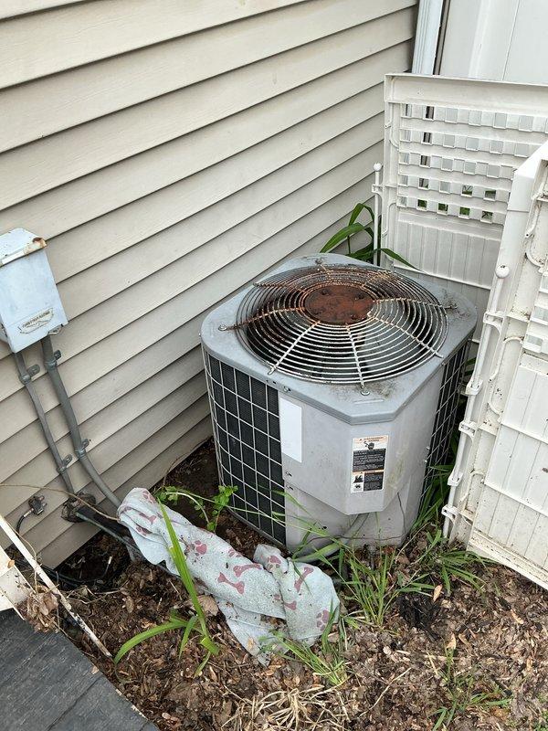 Completed comprehensive Gold AC inspection on residential split system with outdoor condensing unit located at side of property and air handler positioned on main floor. Unit confirmed operational prior to inspection. Performed thorough visual assessment of outdoor condenser, noting weathering and dirt accumulation on fan guard and housing consistent with age of equipment. Inspected refrigerant lines, electrical conduit, and mounting pad for proper installation and condition. Documented equipment positioning between buildings with lattice screening enclosure and vegetation growth around base requiring attention for optimal airflow. Verified manufacturer information plate present and legible on unit. Inspection completed under suitable weather conditions as required for accurate system evaluation.