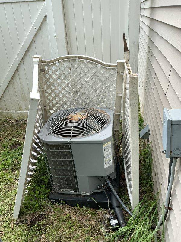Completed comprehensive Gold AC inspection on residential split system with outdoor condensing unit located at side of property and air handler positioned on main floor. Unit confirmed operational prior to inspection. Performed thorough visual assessment of outdoor condenser, noting weathering and dirt accumulation on fan guard and housing consistent with age of equipment. Inspected refrigerant lines, electrical conduit, and mounting pad for proper installation and condition. Documented equipment positioning between buildings with lattice screening enclosure and vegetation growth around base requiring attention for optimal airflow. Verified manufacturer information plate present and legible on unit. Inspection completed under suitable weather conditions as required for accurate system evaluation.
