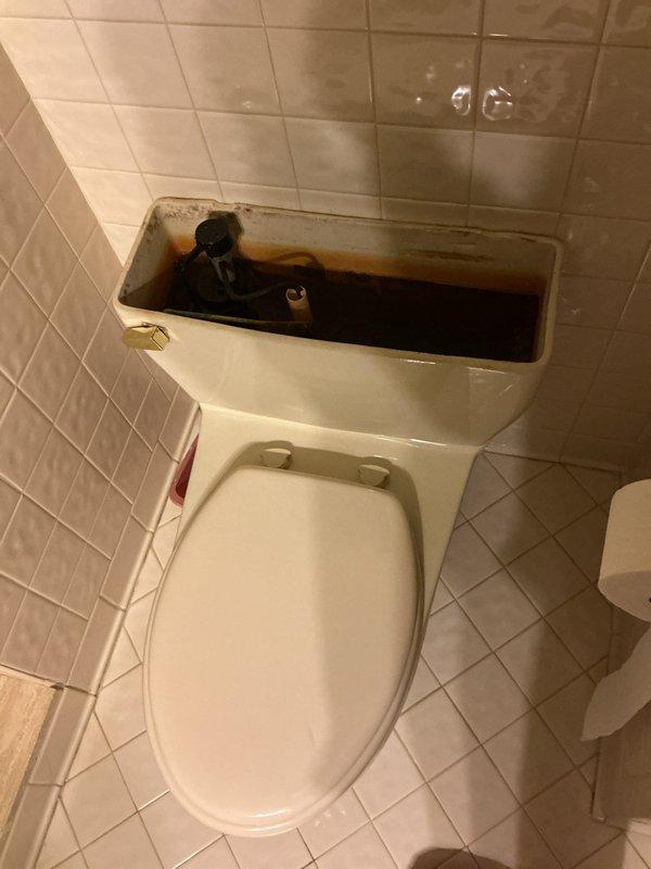 Responded to water leak complaint affecting downstairs ceiling originating from upstairs bathroom. Upon inspection, identified toilet as source of leak. Removed tank lid and discovered significant rust-colored staining and mineral deposits throughout interior tank walls, particularly concentrated at water line. Water inside tank showed visible discoloration indicating corroded internal components and deteriorated tank integrity. Internal flush valve and fill valve mechanisms exhibited signs of age-related wear and corrosion. Leak likely caused by compromised tank-to-bowl connection or deteriorated tank components allowing water seepage through ceiling below. Documented conditions with photographs showing extent of rust accumulation and internal tank degradation requiring component replacement or full toilet replacement to resolve leak issue.