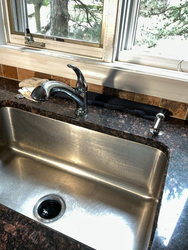Completed installation of waste and overflow assembly for undermount stainless steel kitchen sink with connection to existing copper supply lines and drainage system. Installed chrome single-handle pull-down faucet and coordinating soap dispenser in dark granite countertop. Verified all connections for proper operation and tested system for leaks at basement level plumbing infrastructure where copper supply lines and shutoff valves service the kitchen fixtures above.