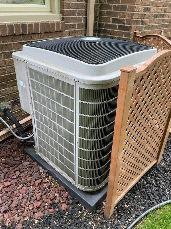 Completed Gold AC Inspection on a 4-year-old residential air conditioning system with outdoor condenser unit and basement-located air handler. Performed comprehensive maintenance evaluation of the outdoor unit positioned on concrete pad against brick exterior, verified proper installation on vibration dampening pad, inspected condenser coils through side grilles for cleanliness and damage, checked electrical connections and disconnect box mounting, and assessed overall system condition. Unit showed proper installation with adequate clearance, decorative lattice screening for aesthetic purposes, and appropriate electrical conduit routing, with all components appearing operational and well-maintained for the equipment age.