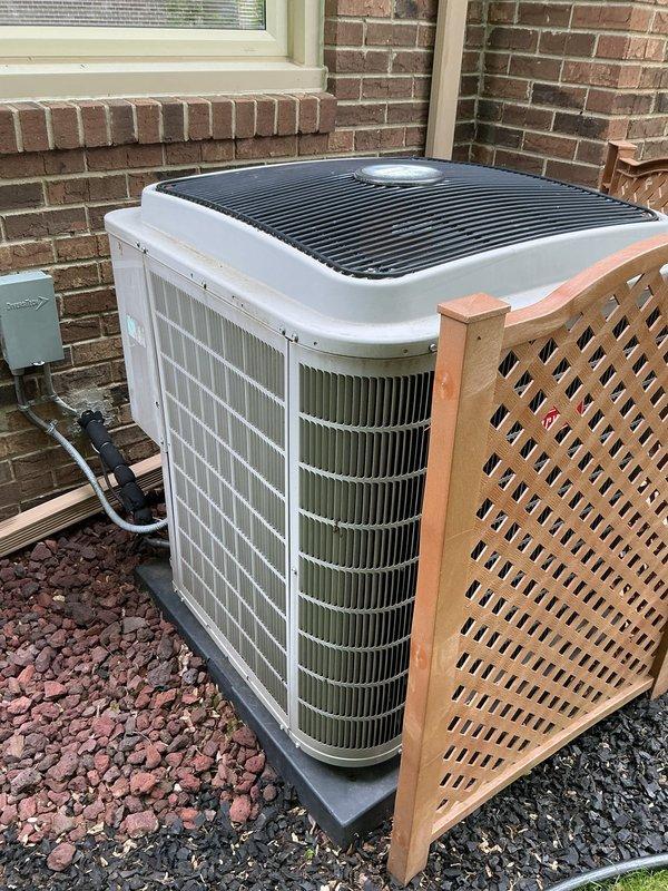 Completed Gold AC Inspection on a 4-year-old residential air conditioning system with outdoor condenser unit and basement-located air handler. Performed comprehensive maintenance evaluation of the outdoor unit positioned on concrete pad against brick exterior, verified proper installation on vibration dampening pad, inspected condenser coils through side grilles for cleanliness and damage, checked electrical connections and disconnect box mounting, and assessed overall system condition. Unit showed proper installation with adequate clearance, decorative lattice screening for aesthetic purposes, and appropriate electrical conduit routing, with all components appearing operational and well-maintained for the equipment age.