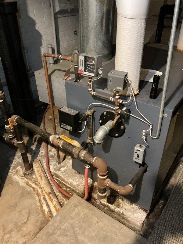 Completed estimate for replacement of 23-year-old steam boiler system currently in operational condition. Unit features gray metal construction with multiple copper supply and return lines, digital control panel, and Honeywell Vaporstat pressure control device housed in protective casing. System includes standard electrical components, brass valving, and various pipe connections utilizing copper fittings and flexible connectors. Equipment is properly installed on concrete floor with wooden base platform in basement utility room with white brick walls and exposed ductwork. All components photographed and documented for replacement proposal.