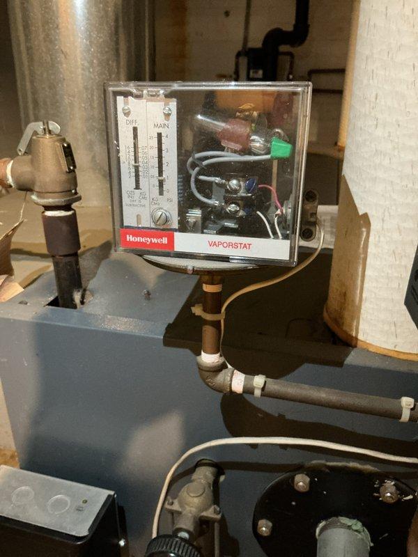 Completed estimate for replacement of 23-year-old steam boiler system currently in operational condition. Unit features gray metal construction with multiple copper supply and return lines, digital control panel, and Honeywell Vaporstat pressure control device housed in protective casing. System includes standard electrical components, brass valving, and various pipe connections utilizing copper fittings and flexible connectors. Equipment is properly installed on concrete floor with wooden base platform in basement utility room with white brick walls and exposed ductwork. All components photographed and documented for replacement proposal.