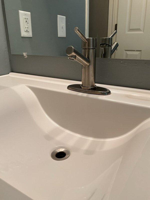 Completed installation of new bathroom sink faucet assembly with modern single-lever pull-out design in brushed nickel finish, including mounting plate and drain hardware. Replaced deteriorating P-trap assembly beneath sink due to visible corrosion and evidence of previous water leakage, installing new white plastic trap with proper sealing connections. Connected chrome braided supply line to ensure proper water flow to new faucet fixture.