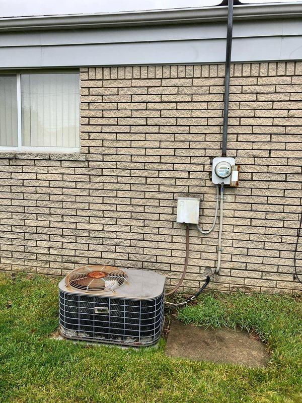 Conducted comprehensive evaluation of 15-22 year old residential air conditioning system experiencing cooling failure. Diagnosed multiple critical issues including low refrigerant charge, blown electrical fuse, and damaged wiring in basement-mounted equipment. System exhibited freezing condition due to compromised cooling capacity. Assessed exterior condensing unit with beige metal casing and copper fan assembly mounted on foundation pad alongside brick wall exterior. Evaluated electrical conduit routing, junction boxes, and power supply connections. Provided detailed replacement estimate for aging HVAC system due to cumulative component failures and overall equipment condition. Documented existing installation configuration including PVC drainage, flexible conduit runs, and utility infrastructure mounting. Recommended full system replacement as most viable solution given equipment age and extent of required repairs.