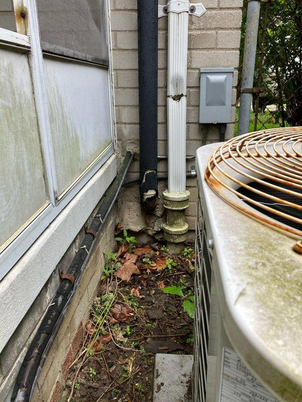 Completed comprehensive air conditioning system evaluation for replacement estimate on residential property with 10+ year old equipment. Air handler and coil currently located in basement. Documented existing outdoor condenser unit showing signs of weathering, algae growth, and debris accumulation in surrounding area including leaves, twigs, and vegetation growth between unit and building foundation. Noted exterior utility infrastructure including electrical panel, meter, and conduit routing along brick exterior wall. Advised property owner that condenser perimeter requires clearance of organic debris and potential animal waste prior to new equipment installation. Generated multiple replacement options and quotes for complete system upgrade.