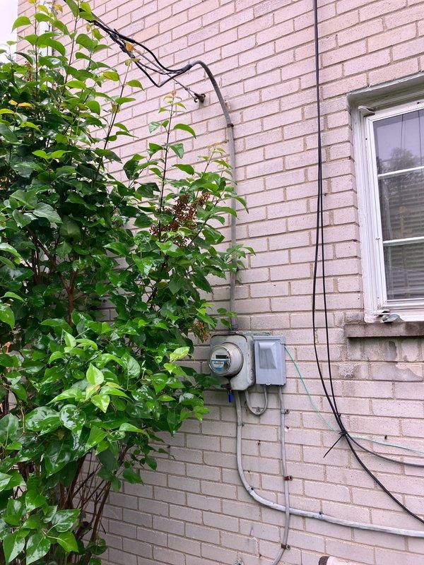 Completed comprehensive air conditioning system evaluation for replacement estimate on residential property with 10+ year old equipment. Air handler and coil currently located in basement. Documented existing outdoor condenser unit showing signs of weathering, algae growth, and debris accumulation in surrounding area including leaves, twigs, and vegetation growth between unit and building foundation. Noted exterior utility infrastructure including electrical panel, meter, and conduit routing along brick exterior wall. Advised property owner that condenser perimeter requires clearance of organic debris and potential animal waste prior to new equipment installation. Generated multiple replacement options and quotes for complete system upgrade.