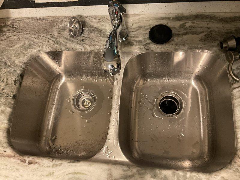 Completed installation of customer-provided kitchen faucet on double-basin stainless steel sink with chrome single-handle fixture mounted centrally on granite countertop. Inspected under-sink plumbing configuration including PVC P-trap assembly, garbage disposal connections, and flexible supply lines. Addressed customer concern regarding drain line leak connected to washing machine; verified all connections secure and functioning properly upon completion.