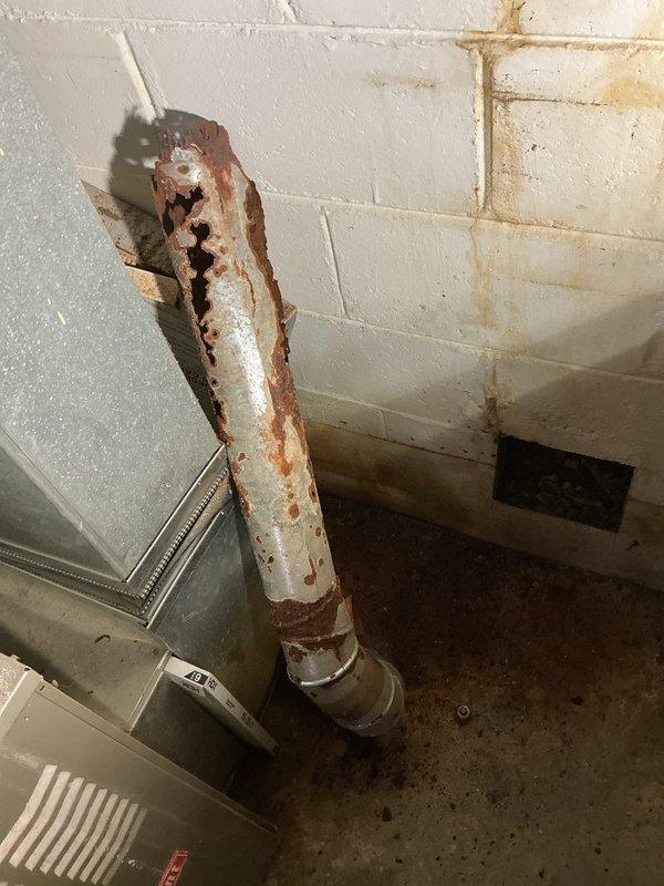 Responded to service call for non-functioning tanked water heater with flame ignition issues. Upon inspection, discovered the exhaust vent pipe above the water heater exhibited severe corrosion and rust deterioration throughout its length. The metal exhaust flue showed extensive oxidation with compromised structural integrity, including visible perforations and rust-through sections. Evidence of moisture infiltration was present at the pipe base with floor staining indicating long-term exposure. The corroded exhaust system posed both a venting hazard and potential carbon monoxide concern, preventing safe water heater operation. Documented the heavily deteriorated condition of the vertical exhaust pipe and surrounding HVAC ductwork, which displayed rust staining on adjacent wall surfaces. The exhaust venting system requires complete replacement to restore safe operation of the water heating appliance and ensure proper combustion gas evacuation from the dwelling.