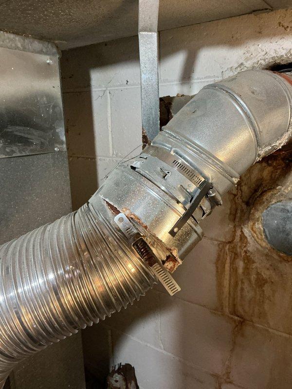 Responded to service call for non-functioning tanked water heater with flame ignition issues. Upon inspection, discovered the exhaust vent pipe above the water heater exhibited severe corrosion and rust deterioration throughout its length. The metal exhaust flue showed extensive oxidation with compromised structural integrity, including visible perforations and rust-through sections. Evidence of moisture infiltration was present at the pipe base with floor staining indicating long-term exposure. The corroded exhaust system posed both a venting hazard and potential carbon monoxide concern, preventing safe water heater operation. Documented the heavily deteriorated condition of the vertical exhaust pipe and surrounding HVAC ductwork, which displayed rust staining on adjacent wall surfaces. The exhaust venting system requires complete replacement to restore safe operation of the water heating appliance and ensure proper combustion gas evacuation from the dwelling.