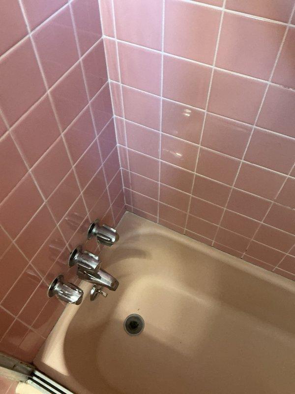 Responded to leaking shower knobs and faucet in main floor bathroom shower-tub combo unit. Upon inspection, accessed plumbing components through wall cavity to assess deteriorated fixtures in 1955-era installation. Evaluated three-handle chrome valve assembly mounted above tub spout showing signs of internal seal failure causing active leak for 2-3 days. Examined condition of supply lines and drain assembly through access point, noting significant age-related wear on mechanical systems. Documented existing pink tile surround and beige tub configuration with original plumbing hardware. Identified source of water seepage at valve stems requiring replacement of internal cartridges or complete valve body assembly to restore proper function and eliminate leaking condition.