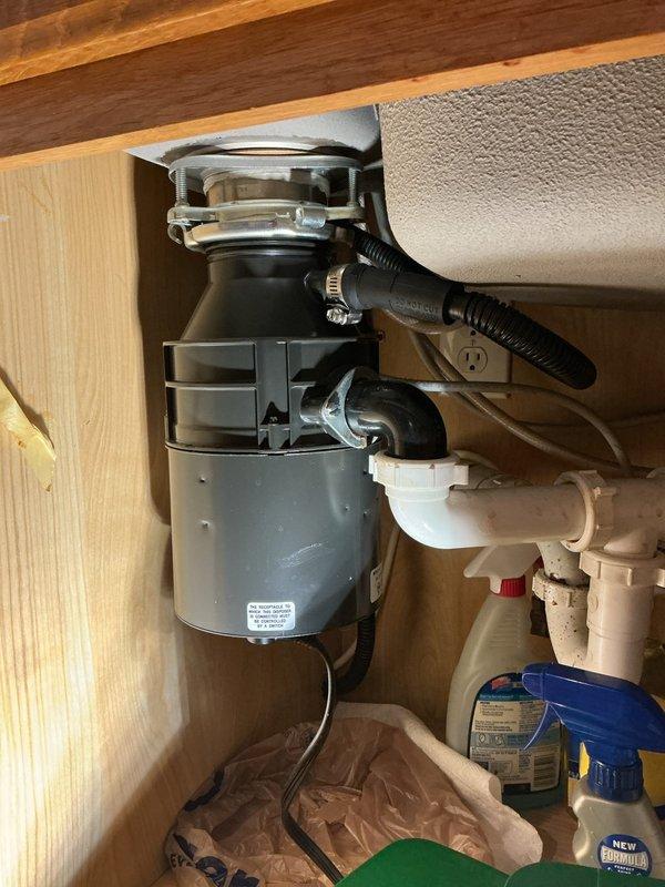 Responded to kitchen plumbing service call for non-functional garbage disposal unit. Upon inspection, determined existing disposal had failed and required complete replacement. Removed old gray disposal unit from mounting assembly beneath sink, disconnected discharge plumbing and electrical connections. Installed new black garbage disposal unit, securing it to existing sink flange with proper mounting hardware. Connected discharge hose to disposal outlet and integrated unit with existing white PVC drain line configuration. Verified electrical connection at cabinet outlet, tested disposal operation for proper function, and confirmed no leaks at mounting flange or drainage connections. Unit operating normally upon completion.