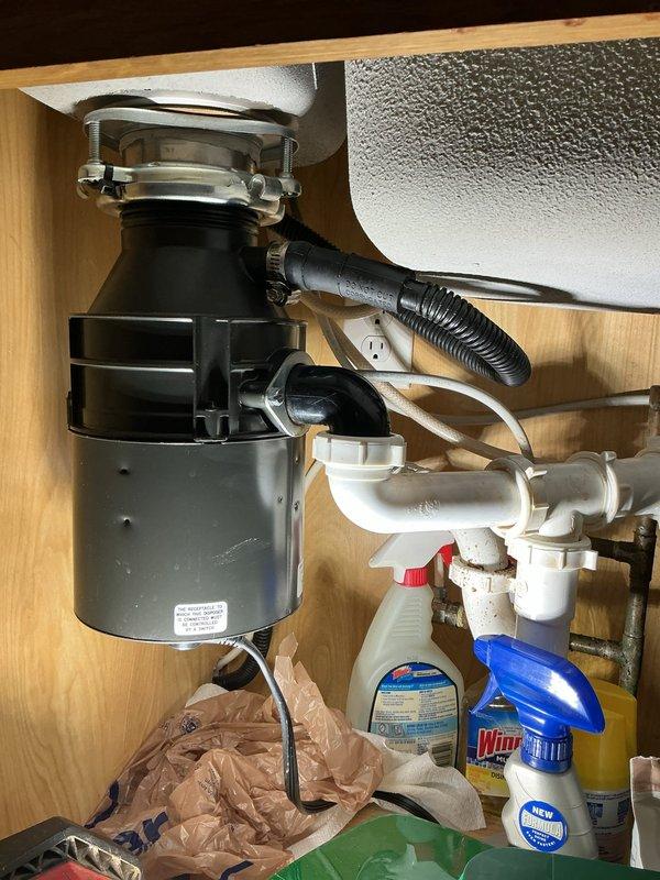 Responded to kitchen plumbing service call for non-functional garbage disposal unit. Upon inspection, determined existing disposal had failed and required complete replacement. Removed old gray disposal unit from mounting assembly beneath sink, disconnected discharge plumbing and electrical connections. Installed new black garbage disposal unit, securing it to existing sink flange with proper mounting hardware. Connected discharge hose to disposal outlet and integrated unit with existing white PVC drain line configuration. Verified electrical connection at cabinet outlet, tested disposal operation for proper function, and confirmed no leaks at mounting flange or drainage connections. Unit operating normally upon completion.