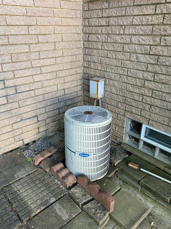 Conducted on-site evaluation of existing Carrier air conditioning system for replacement estimate. Inspected outdoor condenser unit positioned in corner location between brick walls, assessed electrical disconnect box showing weathering and rust staining, and documented current installation configuration including flexible conduit connections. Photographed equipment placement, surrounding patio pavers with moss growth, and mounting setup to provide comprehensive replacement pricing proposal.