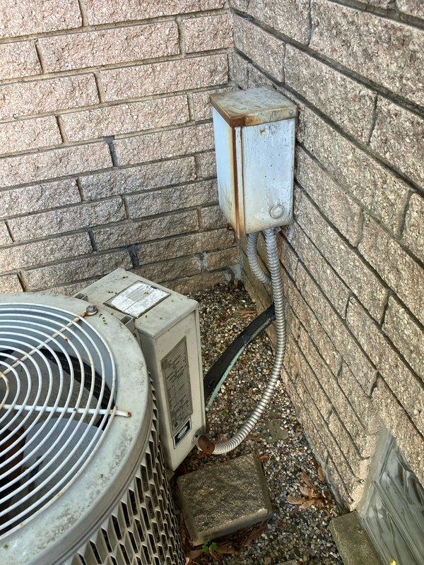 Conducted on-site evaluation of existing Carrier air conditioning system for replacement estimate. Inspected outdoor condenser unit positioned in corner location between brick walls, assessed electrical disconnect box showing weathering and rust staining, and documented current installation configuration including flexible conduit connections. Photographed equipment placement, surrounding patio pavers with moss growth, and mounting setup to provide comprehensive replacement pricing proposal.