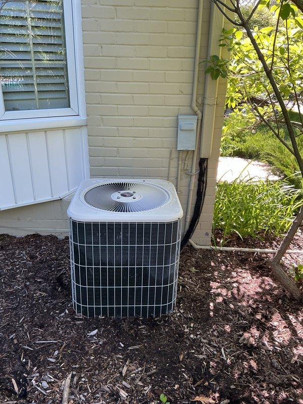 Completed Gold AC inspection on residential property with two HVAC systems: a 25-year-old attic unit utilizing R22 refrigerant and a 12-year-old unit with air handler/coil located in basement. Accessed attic unit using 6-foot ladder to perform comprehensive system evaluation. Documented exterior condensing unit condition through photographic inspection, noting cylindrical condenser configuration with white top cap and metal grille construction, mounted on mulch bed against residential siding with electrical disconnect box and conduit properly installed. Both systems confirmed operational at time of inspection, meeting prerequisites for estimate development on equipment replacement options.