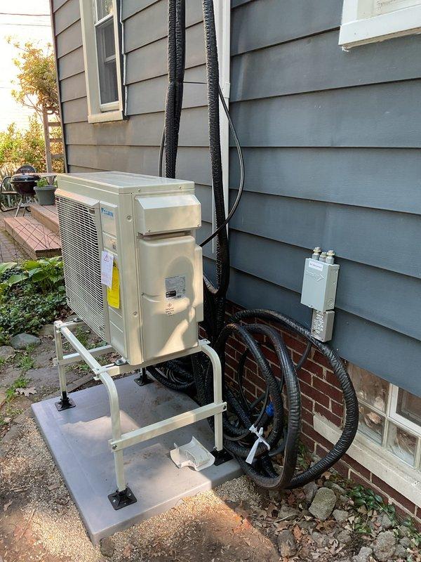 Completed two-day Daikin ductless mini-split system installation including outdoor condenser unit mounted on elevated bracket with concrete pad foundation, refrigerant line sets routed vertically along exterior siding with protective conduit wrapping, electrical disconnect box installation, and interior wall-mounted air handler with proper drainage configuration. System commissioned and tested for proper operation with all components functioning within manufacturer specifications.