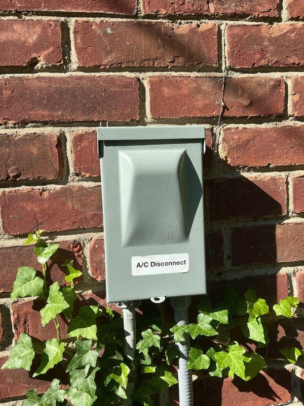 Completed comprehensive Gold AC inspection on 3-year-old single-unit air conditioning system with air handler located in basement. Verified system operational status, inspected outdoor condenser unit positioned on concrete pad with proper clearance, examined electrical disconnect box mounted on exterior brick wall, and confirmed all components functioning within normal parameters. Documented equipment condition through photographic records showing condenser unit, disconnect switch, and surrounding area with minor vegetation growth present but not obstructing airflow or access.