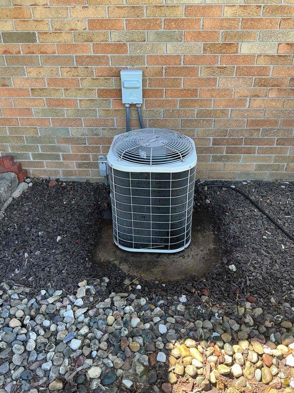 Completed Gold AC inspection on 22-year-old residential air conditioning system with air handler located in basement. Performed comprehensive evaluation of outdoor condenser unit, which is positioned on concrete pad against brick exterior wall with proper electrical disconnect box mounted above. Verified unit operational status, inspected heat exchanger coils, fan assembly, and protective grilles for proper condition and functionality. Documented equipment condition through photographic records showing condenser placement, surrounding landscape materials including decorative rock and soil, and overall system accessibility. Unit appears functional with normal aging characteristics for equipment of this vintage, with condenser properly secured on weathered concrete pad and adequate clearance maintained around perimeter for airflow.