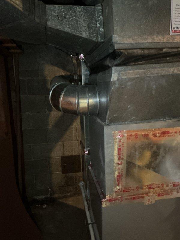Conducted on-site evaluation of existing 32-year-old air conditioning system located on exterior side of residence to provide replacement estimate. Inspected current outdoor condensing unit and assessed overall system condition, noting equipment age has exceeded typical service life expectancy. Evaluated interior HVAC components including ductwork assembly, furnace unit with access panels, and associated mechanical connections in basement utility area. Documented existing system configuration through photographic records showing dark-colored metal ductwork, cylindrical pipe connections, and furnace equipment with transparent inspection panels. Reviewed current installation layout to determine replacement specifications and equipment requirements for new air conditioning system installation.