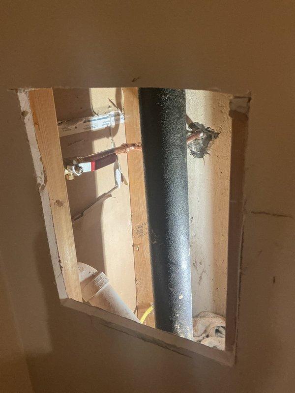 Accessed workshop plumbing system through existing wall opening to inspect and photograph concealed piping infrastructure. Located and documented galvanized ductwork, copper supply lines with shut-off valves (hot and cold), and PVC drainage piping within wall cavity. Verified proper installation and accessibility of plumbing components behind finished drywall surface for maintenance and future service requirements.