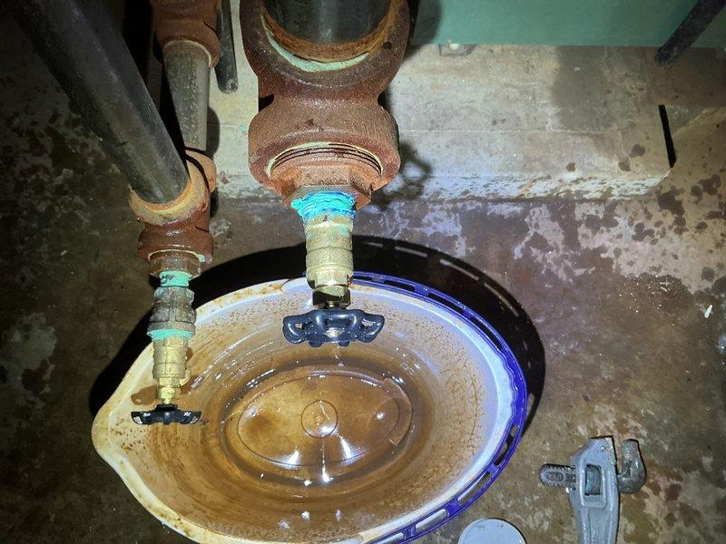 Responded to reported leak near boiler system with uncertainty about source involving multiple drain valves. Upon inspection, identified active leak from one of four boiler drain faucets located in basement utility area serving a 4-year-old boiler system. Found significant corrosion and rust buildup on copper piping and valve fittings, with discolored water present in collection basin indicating ongoing leak and mineral deposits. Observed deteriorated valve components with visible patina on copper connections and staining patterns consistent with intermittent water discharge. Assessed condition of all four drain valves connected to boiler system, noting age-related wear on valve seats and handles. Documented plumbing configuration showing ball valve controls and galvanized connections with rust-colored staining on metal components. Identified mineral buildup in collection area and standing water with sediment accumulation. Photographed existing conditions including corroded valve assem
