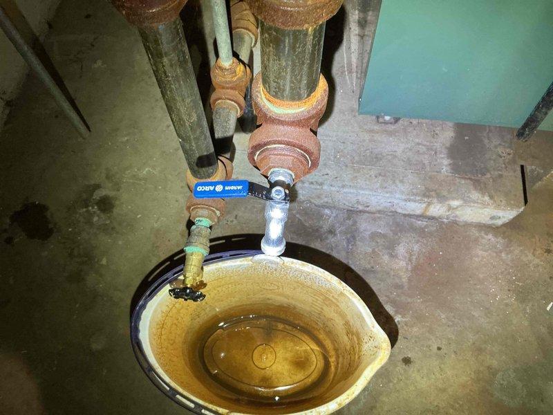 Responded to reported leak near boiler system with uncertainty about source involving multiple drain valves. Upon inspection, identified active leak from one of four boiler drain faucets located in basement utility area serving a 4-year-old boiler system. Found significant corrosion and rust buildup on copper piping and valve fittings, with discolored water present in collection basin indicating ongoing leak and mineral deposits. Observed deteriorated valve components with visible patina on copper connections and staining patterns consistent with intermittent water discharge. Assessed condition of all four drain valves connected to boiler system, noting age-related wear on valve seats and handles. Documented plumbing configuration showing ball valve controls and galvanized connections with rust-colored staining on metal components. Identified mineral buildup in collection area and standing water with sediment accumulation. Photographed existing conditions including corroded valve assem