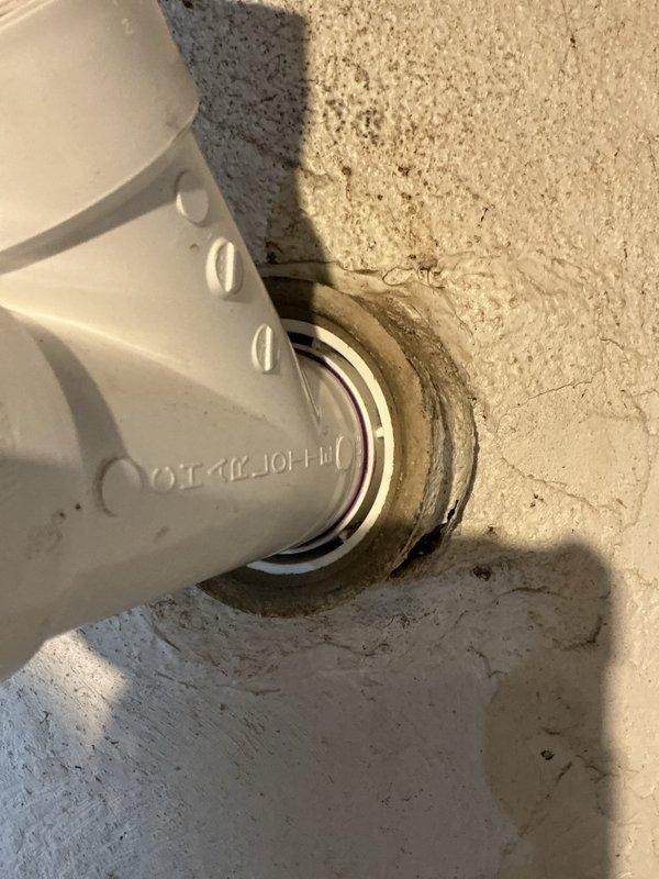Inspected and documented PVC pipe penetration through concrete floor/wall junction. The white PVC conduit with ventilation slots was found to be properly secured with metal compression fittings at the concrete entry point. Verified the integrity of the pipe installation and surrounding concrete surfaces, noting minor debris accumulation that was typical for the location. The pipe and fittings were determined to be in good working condition with no visible damage or signs of failure requiring immediate repair.