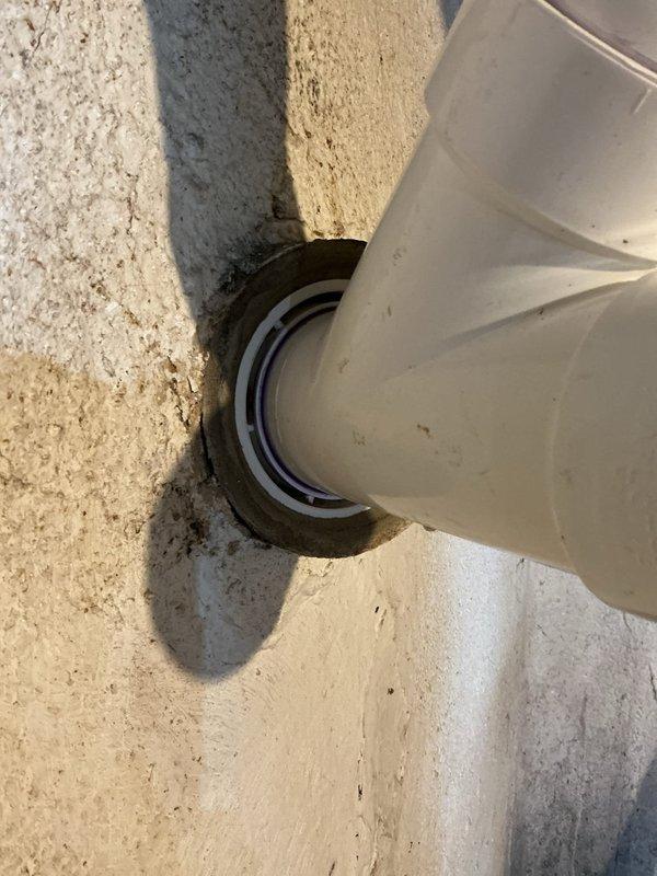 Inspected and documented PVC pipe penetration through concrete floor/wall junction. The white PVC conduit with ventilation slots was found to be properly secured with metal compression fittings at the concrete entry point. Verified the integrity of the pipe installation and surrounding concrete surfaces, noting minor debris accumulation that was typical for the location. The pipe and fittings were determined to be in good working condition with no visible damage or signs of failure requiring immediate repair.