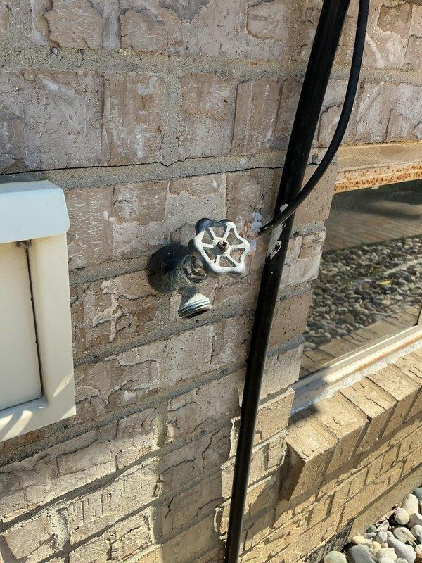 Replaced outdoor hose bib mounted on exterior concrete block wall with new chrome wheel-handle valve assembly. Installed corresponding shut-off valve with blue handle in ceiling joist area above spigot location, connecting to existing copper supply line to provide interior water control for the outdoor fixture.