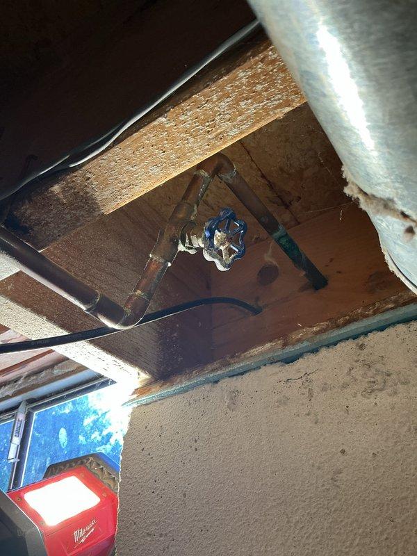 Replaced outdoor hose bib mounted on exterior concrete block wall with new chrome wheel-handle valve assembly. Installed corresponding shut-off valve with blue handle in ceiling joist area above spigot location, connecting to existing copper supply line to provide interior water control for the outdoor fixture.