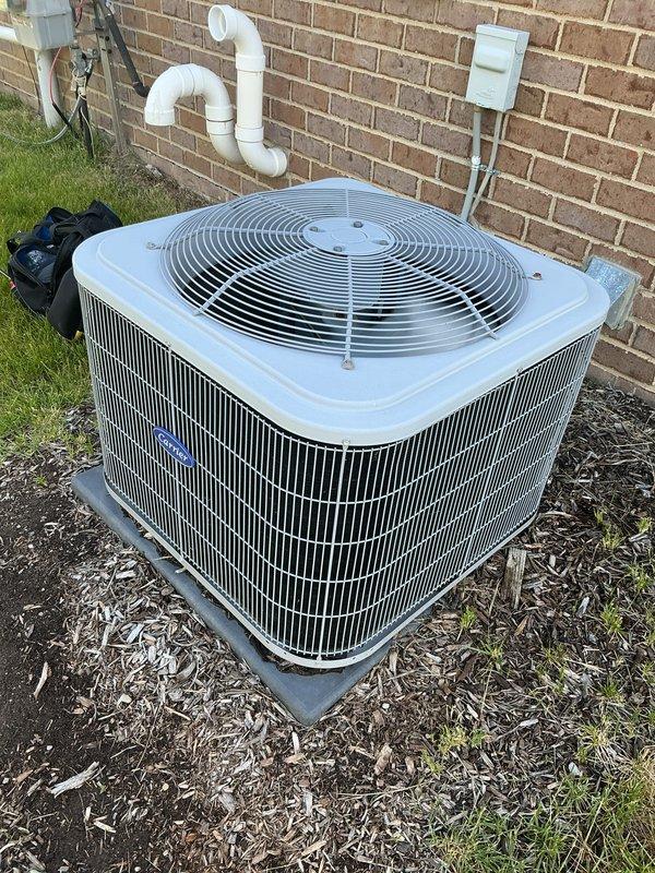 Completed Gold AC Inspection on a 6-year-old air conditioning system with condenser unit located on exterior concrete pad against brick wall and evaporator coil in basement. Performed comprehensive maintenance including visual inspection of outdoor condenser unit, verified proper positioning on concrete pad with adequate clearance, inspected cabinet housing and protective grilles for structural integrity, and confirmed fan guard assembly secure. Checked condensate drainage system including white PVC piping configuration mounted to exterior wall, verified electrical disconnect box properly installed and accessible. Inspected condenser coil fins through louvers, assessed unit for signs of wear, corrosion, or damage. Evaluated surrounding area including mulch bedding and confirmed no debris obstruction to airflow. Unit operating within normal parameters with clean metal surfaces and no visible deficiencies noted. System demonstrates proper installation standards and maintenance condition 